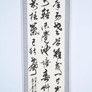 Könnte beinhalten: Eine traditionelle japanische Kalligraphie-Rolle mit schwarzer Tinte auf weißem Papier. Die Rolle zeigt eine Reihe von Schriftzeichen, die in einem fließenden, eleganten Stil geschrieben sind. Die Rolle ist auf einem hellblauen Stoff mit Blumenmuster montiert.