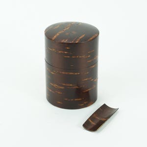 May include: A dark brown wooden tea caddy with a matching lid and a small, curved scoop. The caddy is cylindrical with a glossy finish and a natural wood grain pattern. The scoop is placed beside the caddy.