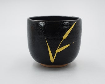 May include: A black ceramic teacup with a gold leaf design. The cup has a rounded shape and a slightly flared rim.