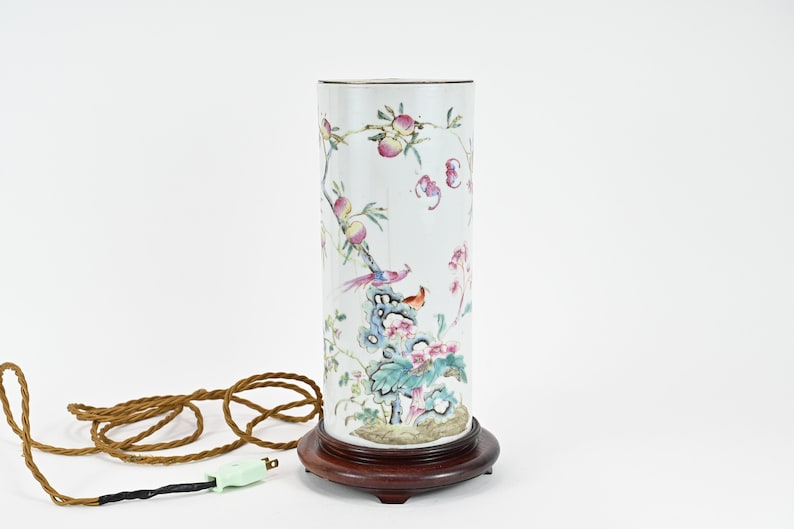 K&ouml;nnte beinhalten: Eine wei&szlig;e Porzellan-Lampenbasis mit einem floralen und Vogelmotiv. Die Basis ist zylindrisch und steht auf einem dunklen Holzsockel. Die Lampe hat ein braunes Kabel und einen gr&uuml;nen Stecker.