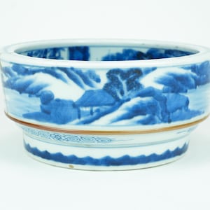 Op de afbeelding: Een ronde, wit en blauw keramische kom met een landschapsontwerp. De kom heeft een blauw-witte berg- en boomscène rond de buitenkant. De kom heeft een bruine band en een decoratief patroon bij de basis.