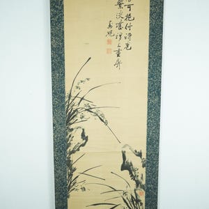 May include: A vertical scroll with an East Asian ink painting. The artwork shows stylized plants and rocks in black ink on a cream background. Calligraphy is at the top. The scroll has a dark blue patterned border. The artwork is a traditional style.