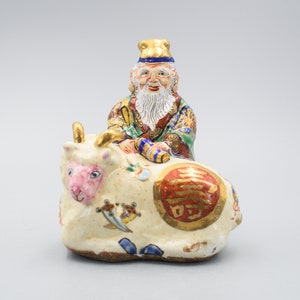Könnte beinhalten: Eine weiße Keramikfigur eines Mannes mit langem weißem Bart, der auf einem weißen Ochsen sitzt. Der Mann trägt eine bunte Robe und eine goldene Krone. Der Ochse hat eine rosa Nase und ist mit goldenen und roten Details verziert. Die Figur ist ein traditionelles chinesisches Symbol für Glück und Wohlstand.