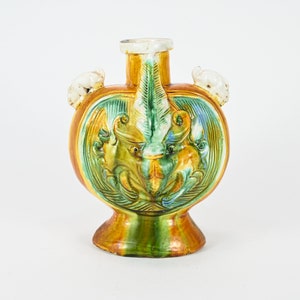 May include: A heart-shaped ceramic vase with green, yellow, and brown glaze. The vase has two small handles on the sides and a detailed design of two mythical creatures in the center.