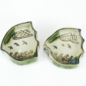 May include: Two ceramic, boat-shaped dishes. Each dish has a light green rim, a cream-colored interior with black bird and wave designs, and a dark brown and blue accent. The dishes are placed on a white background.