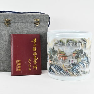 Op de afbeelding: Een witte porseleinen cilinder met een geschilderd landschapsscene van een Chinees dorp met bergen en bomen. De cilinder bevindt zich in een grijze doos met een rood certificaat waarop "Certificaat" en "Chinese antieke kunst" staat.