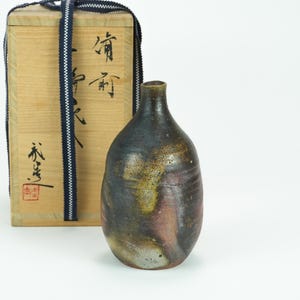 Puede incluir: Una botella de cerámica oscura y moteada con cuello estrecho, junto a una caja de madera con caligrafía japonesa y un cordón azul y blanco. La botella tiene una forma única y una mezcla de tonos marrones, negros y rosas.