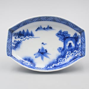 Puede incluir: Un plato de porcelana azul y blanco con un diseño de paisaje. El plato tiene forma ovalada y un borde festoneado. El diseño presenta montañas, árboles y un barco.