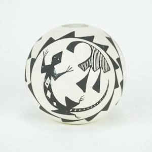 Könnte beinhalten: Eine weiße Keramikvase mit einem schwarzen geometrischen Design mit zwei stilisierten Eidechsen. Die Vase hat eine runde Form und eine kleine Öffnung oben. Das Design ist symmetrisch und bedeckt die gesamte Oberfläche.