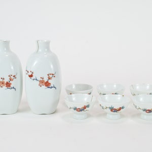 May include: A set of white ceramic sake bottles and cups with a floral design. The bottles and cups have a delicate floral pattern in shades of red, blue, and green.