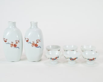 May include: A set of white ceramic sake bottles and cups with a floral design. The bottles and cups have a delicate floral pattern in shades of red, blue, and green.