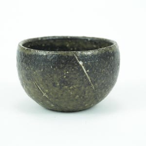 May include: A small, round, handmade ceramic bowl with a dark green and speckled finish. The bowl has a textured surface and a diagonal cut across the side. The interior is dark, and the bowl sits on a white background.
