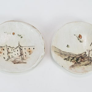 Può includere: Due piccole ciotole in ceramica bianca con scene dipinte a mano di edifici e paesaggi. Le ciotole hanno un aspetto rustico e fatto a mano e presentano una tavolozza di colori marrone chiaro e arancione.