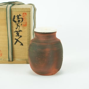 May include: A small, handcrafted, reddish-brown ceramic jar with a white lid. The jar is next to a wooden box with Japanese calligraphy and a woven strap. The box is light-coloured with dark text and red seals.