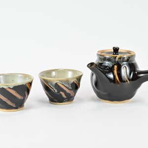 以下が含まれることがあります： 茶色の釉薬がかかった黒い陶器の急須と、それに合わせた2つの茶碗のセット。急須は短い注ぎ口と持ち手があります。茶碗は小さく丸いです。