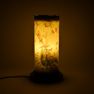 K&ouml;nnte beinhalten: Eine zylindrische Lampe mit einem wei&szlig;en Sockel und einem klaren Glasschirm. Der Schirm ist mit einem Blumenmuster und einem Vogelmotiv verziert. Die Lampe ist eingeschaltet und strahlt ein warmes Licht aus.