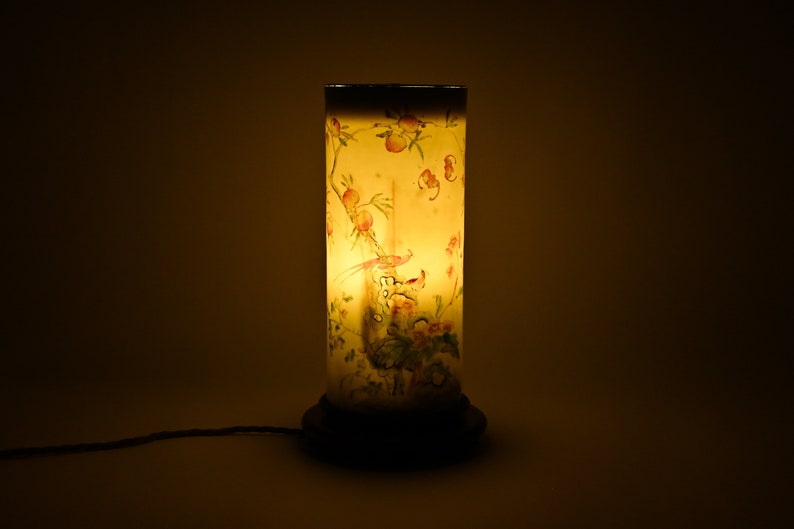 K&ouml;nnte beinhalten: Eine zylindrische Lampe mit einem wei&szlig;en Sockel und einem klaren Glasschirm. Der Schirm ist mit einem Blumenmuster und einem Vogelmotiv verziert. Die Lampe ist eingeschaltet und strahlt ein warmes Licht aus.