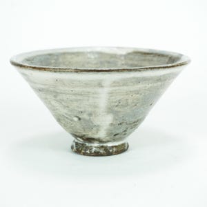 Puede incluir: Un cuenco de cerámica hecho a mano con forma cónica y ancha. El cuenco tiene un esmalte moteado gris claro y blanco, un borde y una base de color marrón oscuro. Una pieza de arte funcional única.