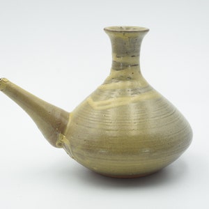 Puede incluir: Una jarra de cerámica verde con un pico largo y un cuerpo redondeado. La jarra tiene un esmalte marrón claro con un sutil patrón de rayas.