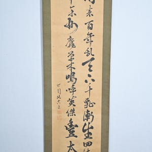 Könnte beinhalten: Ein traditionelles japanisches Rollbild mit schwarzer Tintenkalligraphie auf beigefarbenem Hintergrund. Die Rolle ist auf einem grünen Stoff mit goldenen Akzenten montiert. Der Text lautet "雨素百年乱て八千嘉南生田伤后 六七ふわ 魔る木鳴呼英傑重太阁".