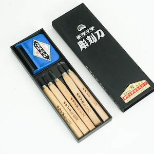 Puede incluir: Un juego de herramientas para tallar madera en una caja negra. El juego incluye varios cuchillos para tallar con mangos de madera y una herramienta de afilado azul. La caja tiene texto japonés.