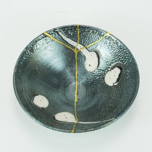 May include: A dark gray ceramic bowl with a textured surface and white accents. The bowl features gold-colored kintsugi repair lines. The bowl is round and has a unique, handcrafted appearance.