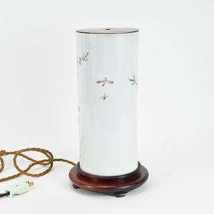 K&ouml;nnte beinhalten: Wei&szlig;e Porzellanlampe mit Blumenmuster und Holzsockel. Die Lampe hat ein braunes Kabel und einen gr&uuml;nen Stecker.