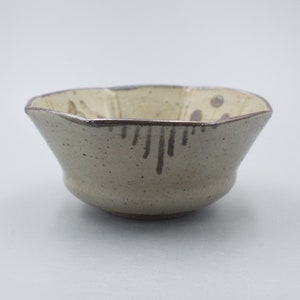 Può includere: Una piccola ciotola in ceramica marrone chiaro con una forma leggermente ottagonale irregolare. La ciotola ha una glassa macchiettata con accenti marroni e una glassa marrone più scura intorno al bordo.