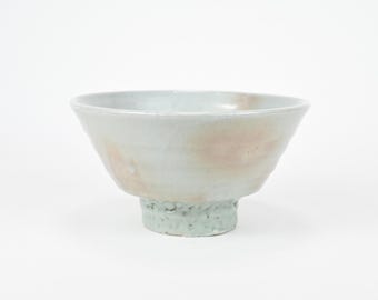 May include: A ceramic bowl with a wide, slightly flared rim and a footed base. The bowl has a light grey glaze with subtle pink and beige tones. The foot is a textured, pale green colour.
