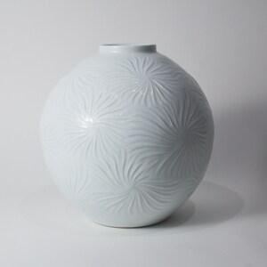 May include: A white ceramic vase with a round body and a textured surface. The vase has a floral design.
