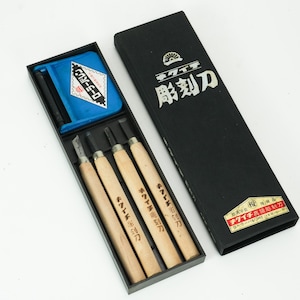 Può includere: Un set di strumenti per intaglio del legno in una scatola nera. La scatola contiene quattro coltelli per intaglio del legno con manici in legno, un panno blu e uno strumento nero. La scatola ha testo giapponese sul coperchio.