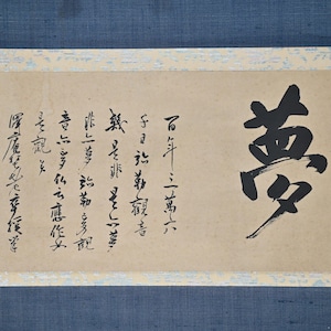 May include: A traditional Japanese scroll painting with black ink calligraphy on a beige background. The scroll features a large character in the centre, surrounded by smaller characters on the left side. The scroll is mounted on a blue fabric backing.