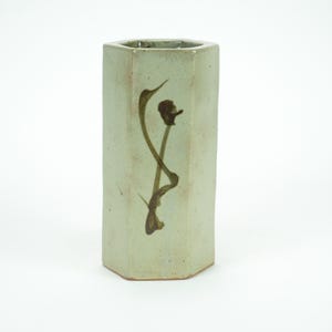 Könnte beinhalten: Eine hohe, sechseckige Vase in einem blassen Grünton. Die Keramikvase hat ein braunes, abstraktes Blumendesign auf der Vorderseite. Die Vase ist ein dekorativer Artikel, der sich zum Ausstellen von Blumen oder als eigenständiges Kunstwerk eignet.