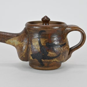 May include: A small brown ceramic teapot with a black and brown abstract design. The teapot has a spout and a handle.