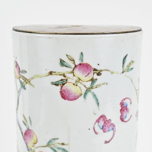 K&ouml;nnte beinhalten: Wei&szlig;e Porzellan-Zylinder-Vase mit Messingdeckel. Die Vase ist mit einem floralen Muster aus rosa und gr&uuml;nen Pfirsichen und Bl&auml;ttern verziert.