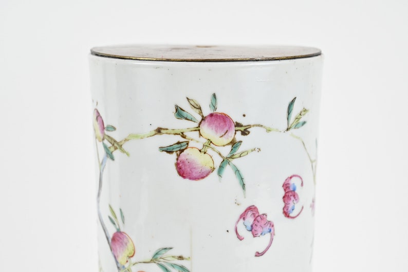 K&ouml;nnte beinhalten: Wei&szlig;e Porzellan-Zylinder-Vase mit Messingdeckel. Die Vase ist mit einem floralen Muster aus rosa und gr&uuml;nen Pfirsichen und Bl&auml;ttern verziert.