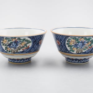 Könnte beinhalten: Zwei kleine, weiße Porzellanschalen mit blauen und goldenen Malereien. Die Schalen zeigen einen Vogel, der auf einem Ast mit Blumen und Blättern sitzt. Die Schalen haben ein blaues und weißes geometrisches Muster auf der Außenseite.