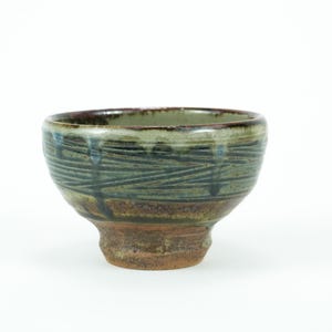 Könnte beinhalten: Eine handgefertigte Keramikschale mit Fuß. Die Schale hat eine strukturierte, glasierte Oberfläche in Grün-, Blau- und Brauntönen. Das Design umfasst horizontale und vertikale Linien, die ein einzigartiges Muster bilden. Ideal zum Servieren oder Ausstellen.