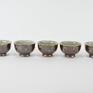 Peut inclure: Cinq petits bols en céramique brune avec un glaçage vert clair. Les bols sont disposés en rangée sur une surface blanche.
