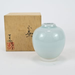 Pode incluir: Um pequeno vaso de cerâmica azul claro com corpo arredondado e base branca. O vaso está sobre uma superfície branca e na frente de uma caixa de madeira com escrita japonesa.