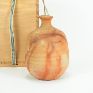 Puede incluir: Una botella de cerámica con cuello estrecho y cuerpo redondeado, con una combinación de colores moteados naranja y marrón. La botella está junto a una caja de madera con una cinta a rayas turquesa y blanca.