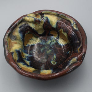 May include: A small, round ceramic bowl with a dark brown, blue, and yellow glaze. The bowl has an uneven, textured surface and a slightly raised rim.