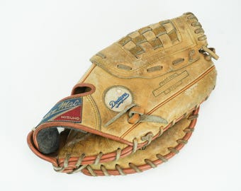 Könnte beinhalten: Ein beigefarbener Baseballhandschuh aus Leder mit einem Dodgers-Teamlogo-Aufnäher, einem Mizuno Air Mac-Etikett mit roten und blauen Akzenten sowie einer dicken Schnürung an den Rändern. Der Handschuh weist Gebrauchsspuren und dunkle Lederstellen auf.