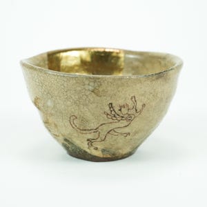 Könnte beinhalten: Keramikschale mit strukturierter, cremefarbener Außenseite und goldfarbenem Innenraum. Auf der Seite befindet sich eine braune Illustration einer Fabelwesen. Die Schale hat eine unregelmäßige Form und ist wahrscheinlich handgefertigt.