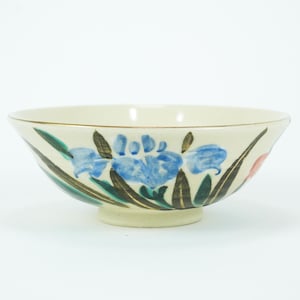 Puede incluir: Cuenco de cerámica color crema con diseño floral. Presenta flores de iris azules, hojas verdes y un borde dorado. El cuenco está sobre una base pequeña y redonda.