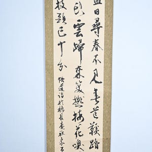 Könnte beinhalten: Ein traditionelles japanisches Rollbild mit schwarzer Tintenkalligraphie auf beigefarbenem Hintergrund. Die Schriftrolle zeigt eine Reihe vertikaler Textzeilen in fließender Schrift. Der Text ist in Japanisch und lautet: "Die Blumen blühen, die Vögel singen und die Menschen sind glücklich."