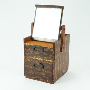 Puede incluir: Un tocador de madera vintage con espejo rectangular y dos cajones. El tocador es de madera marrón oscuro con vetas visibles y tiradores negros. El espejo está sostenido por brazos de madera.