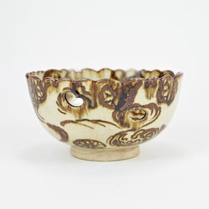 Puede incluir: Un cuenco de cerámica blanca con un diseño floral marrón y un borde festoneado. El cuenco tiene varios agujeros pequeños alrededor del borde.