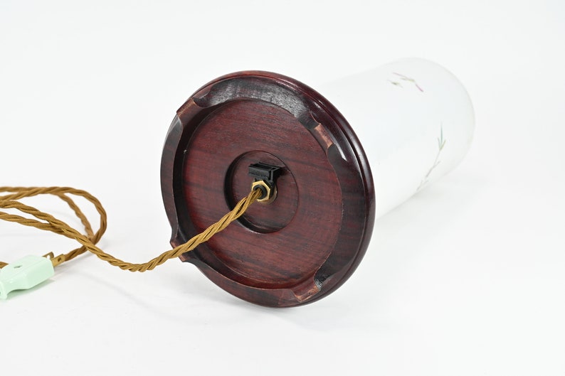 K&ouml;nnte beinhalten: Eine wei&szlig;e Glastischlampe mit einem braunen Holzsockel und einem goldenen Kabel. Die Lampe hat ein schlichtes Design mit einem wei&szlig;en Blumenmuster auf dem Glas.