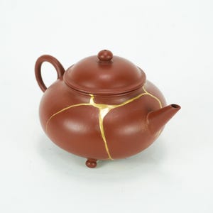 May include: A brown ceramic teapot with a lid, spout, and handle. The teapot has gold-colored lines across its surface, indicating the Kintsugi repair technique. The teapot sits on three small feet.